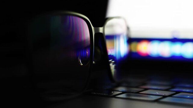 Glasses rest on a keyboard, reflecting bright screen glow. Dark room contrasts with vibrant laptop display colors. Fingers unseen, focus on screen, eyes tired from work. Perfect for tech fatigue