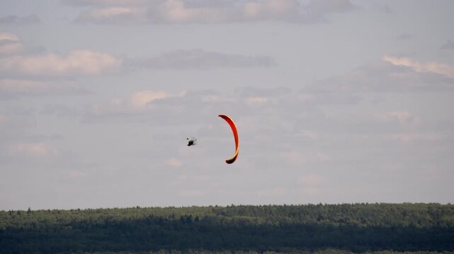 Red paraglider soars above dense green forest. Sky holds soft clouds, sun glows gently. Parachutist glides calmly, enjoying open air. Perfect for adventure, nature, or outdoor sports visuals