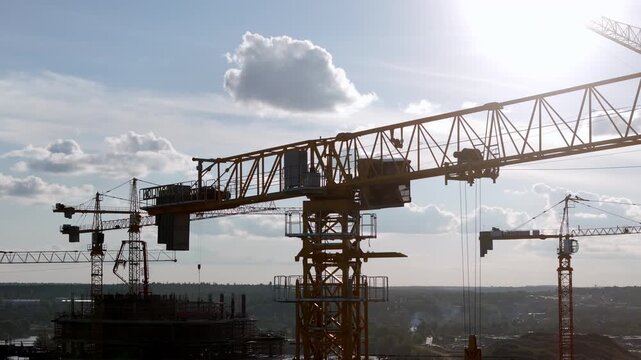 Large cranes dominate the construction skyline. Sunlight casts long shadows across the site. Workers move among the steel structures. Busy development zone under clear skies