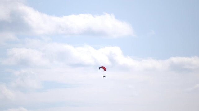 Parachutist glides through soft, blue sky. Clouds drift lazily overhead. Red canopy contrasts gentle daylight. Skydiver soars with calm, focused grace. Perfect for adventure, travel, or freedom themes
