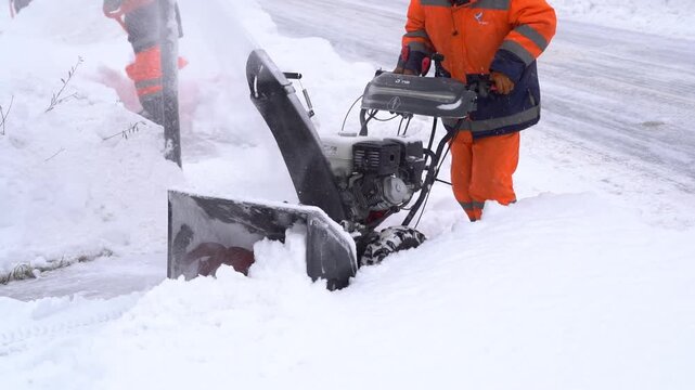 Worker clears snow with snowblower on icy road. Bright orange gear stands out against white snow. Snowblower kicks up powder as it moves forward. Worker's focused effort clears path for safe travel