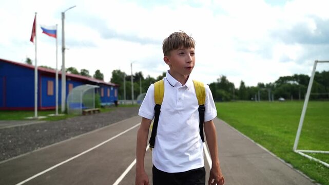 Boy walks calmly on a school track. He wears a white shirt and yellow backpack. His expression is curious and focused. Flags and blue buildings frame the background. Ideal for school, sports