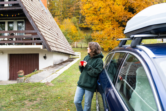 A woman leaning against a blue car with a red mug in her hand in front of a wooden A-frame house surrounded by colorful autumn forest