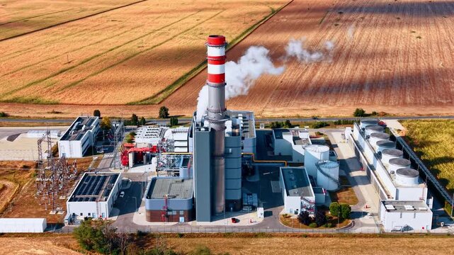 Aerial view of a power station chimney and cooling equipment. A red and white stack rises beside gray structures, storage tanks, fan rows and a fenced electrical yard.