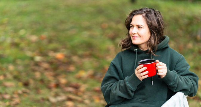 A woman sitting in a wooden chair with a blanket and a red mug, relaxing outdoors during autumn in a countryside garden