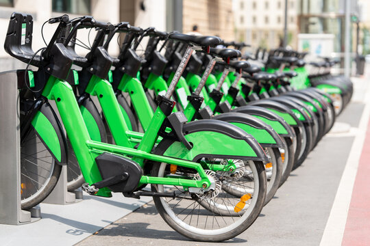 A row of green electric bicycles parked at an urban public bikeshare docking station on a city street.