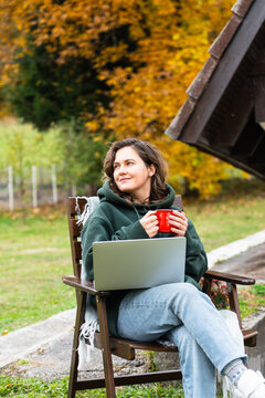 A woman sitting in a wooden chair outside an A-frame house with a laptop and a red mug during autumn