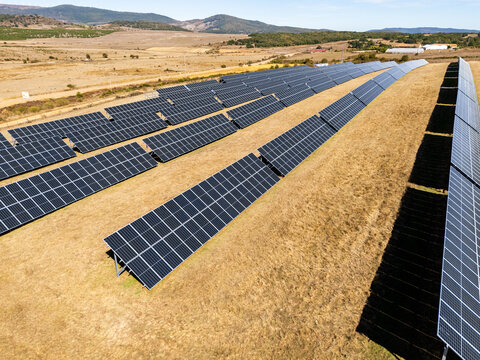 A wide aerial view of a large solar panel farm installed in a dry field under a clear blue sky with mountains in the background