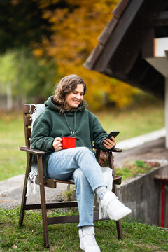 A woman sitting in a wooden chair outside an A-frame house holding a smartphone and a red mug during autumn