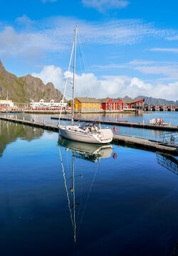 Sailboat at a pier in Svolvaer harbor in the Lofoten Islands with calm water, colorful houses and distinctive mountains beneath a clear blue sky.