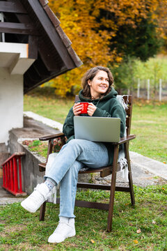 A woman sitting in a wooden chair outside an A-frame house with a laptop and a red mug during autumn