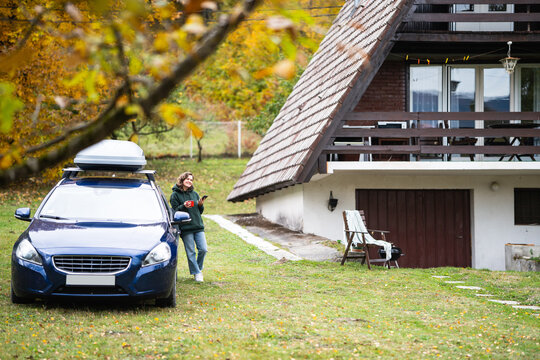 A smiling woman standing next to a blue car with a roof box, holding a red mug and using a smartphone in front of an A-frame house in autumn