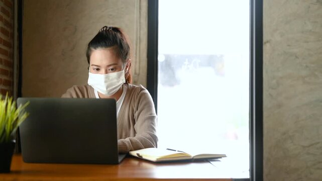 A woman wearing a face mask types on a laptop at a cafe workspace. The woman works beside a notebook and food plate on a wooden table, with window light falling across the brick wall behind her.