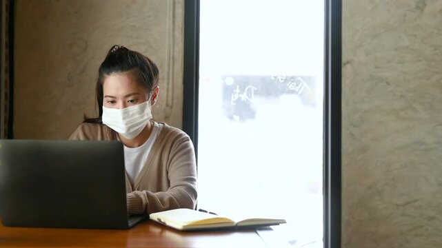 A woman wearing a face mask types on a laptop at a cafe workspace. The woman works beside a notebook and food plate on a wooden table, with window light falling across the brick wall behind her.