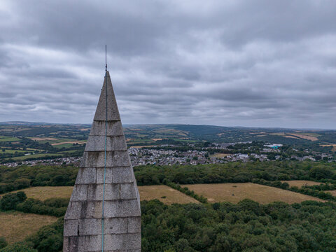 Aerial view of the stone obelisk of Bodmin Beacon rising above surrounding lush green fields, dense woodland, and the town of Bodmin under a cloudy sky. Bodmin, Cornwall, United Kingdom.