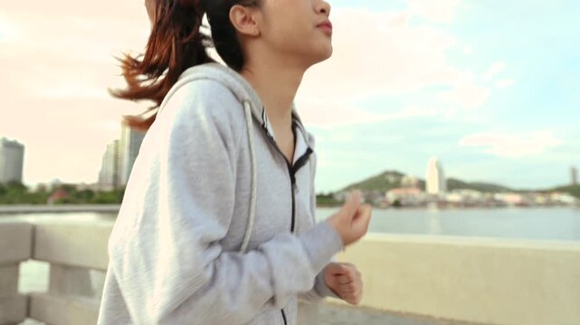 A woman is jogging along a waterfront path in sportswear, with bent arms and a steady running stride. The woman passes a railing while water and distant buildings stretch behind the pavement.