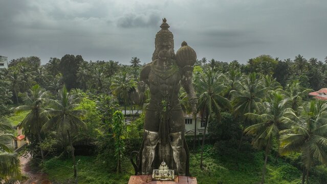 Aerial view of the large Hanuman statue surrounded by lush palm trees and greenery under a cloudy sky in Udupi, Karnataka, India.