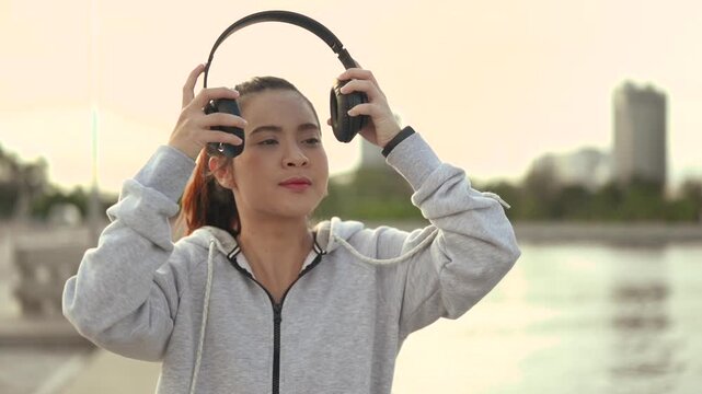 A woman in sportswear stands by the waterfront wearing a gray zip hoodie and athletic leggings. The woman faces the camera near a metal railing, with water and distant city buildings behind her.