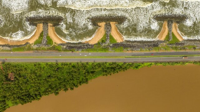 Aerial view of Maravanthe beach featuring unique T-shaped stone breakwaters, crashing waves, a coastal highway running parallel to the shore, and a calm river in Maravanthe, Karnataka, India.