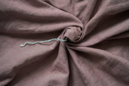 Loose Mint String Extends Left From Small Opening In Gathered Mauve Textile, Surrounded By Deep Curved Folds And Shadowed Ridges Across Fabric Background.