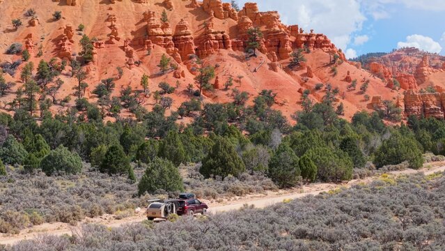 Aerial view of an SUV pulling a teardrop camper trailer along a dirt road through the red rock formations and pine trees of Costa Canyon, Panguitch, Utah, United States.