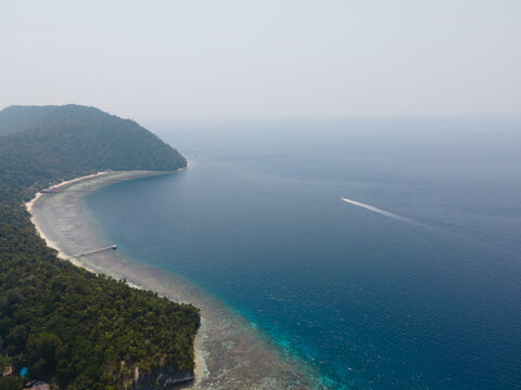 Aerial view of Kri Island with its lush tropical forest, white sandy beach, and a small boat leaving a wake in the clear blue sea in Raja Ampat, Southwest Papua, Indonesia.