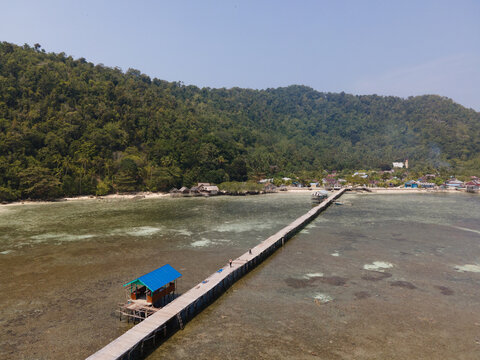 Aerial view of Yenbuba jetty extending into shallow turquoise waters, with lush tropical hills and a small village in the background, Raja Ampat, Southwest Papua, Indonesia.