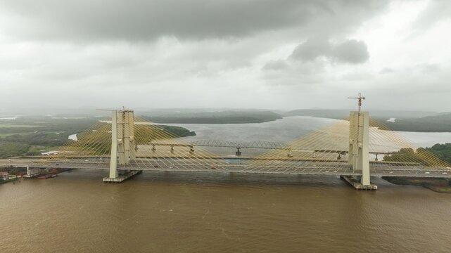 Aerial view of the Zuari Bridge spanning the river with its cable-stayed design, construction cranes, and cloudy sky in the background in Cortalim, Goa, India.