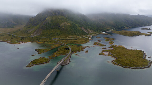 Aerial view of Lofoten bridge connecting islands surrounded by turquoise water, lush green landscape, and dramatic mountains under a cloudy sky in Nordland, Norway.