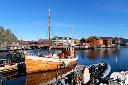 Traditional wooden boat in Svolvaer harbor in the Lofoten Islands with other boats, colorful houses, calm water and Nordic architecture beneath blue sky.
