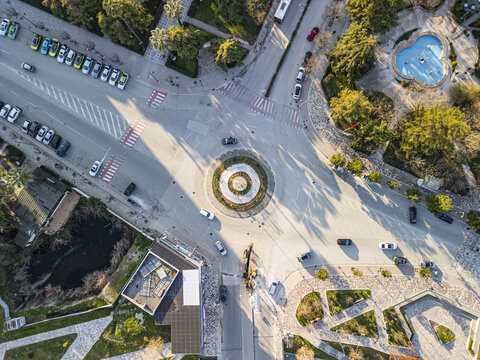 Aerial view of Sheshi Pavaresia featuring a central roundabout, surrounding streets with parked cars, a blue fountain, and urban greenery in Fier, Qarku i Fierit, Albania.