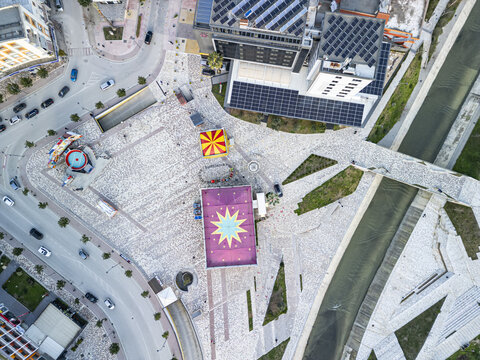 Aerial view of Fier Plaza featuring a patterned stone square, colorful structures, nearby urban streets, and a river flowing alongside in Fier, Fier County, Albania.
