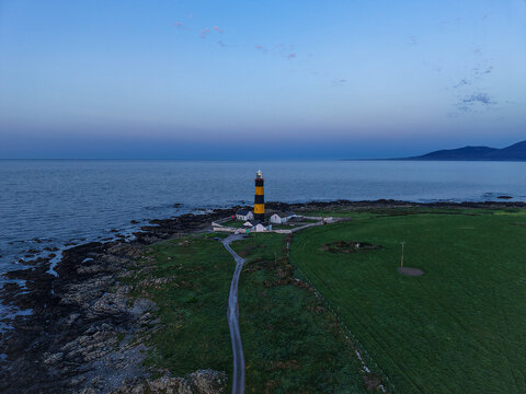 Aerial view of St. John's Point Lighthouse with its distinctive black and yellow stripes, situated on the rocky coastline near green fields in Newcastle, Northern Ireland, United Kingdom.