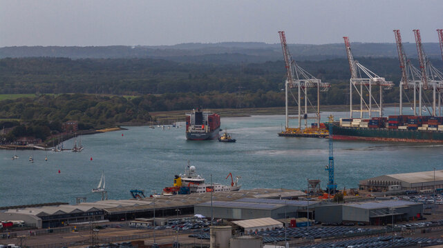 Aerial view of the Port of Southampton with a large container ship being guided by a tugboat, surrounded by industrial cranes and shipping containers in Southampton, England, United Kingdom.