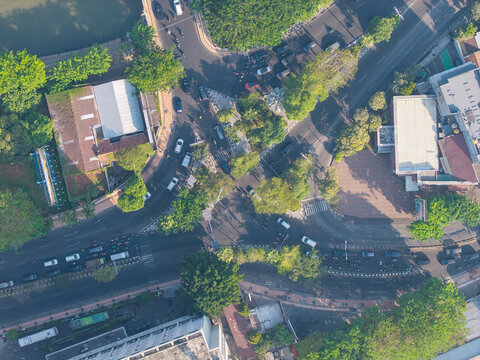 Aerial view of a busy road intersection with cars and motorcycles navigating the streets surrounded by lush green trees and urban buildings in Surabaya, East Java, Indonesia.