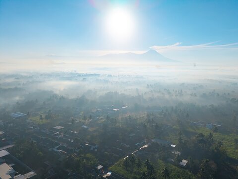 Aerial view of the town of Kepanjen nestled in a valley with morning mist, lush greenery, and a distant mountain under a bright sun in Kepanjen, East Java, Indonesia.