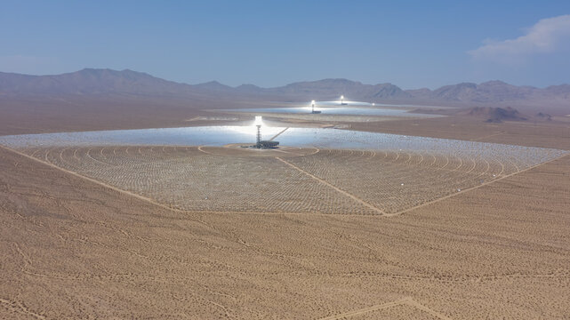 Aerial view of a large concentrated solar power plant with thousands of heliostat mirrors reflecting sunlight toward central solar towers in a vast desert landscape, United States.