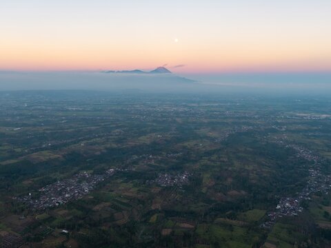 Aerial view of the landscape near Malang with a distant volcano rising above clouds during sunset, featuring scattered settlements and agricultural fields in Malang, East Java, Indonesia.