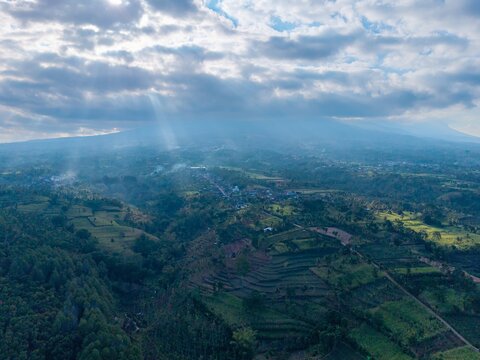 Aerial view of lush terraced fields and rolling hills in Malang, with dramatic sunlight filtering through clouds over distant mountains, Malang, East Java, Indonesia.