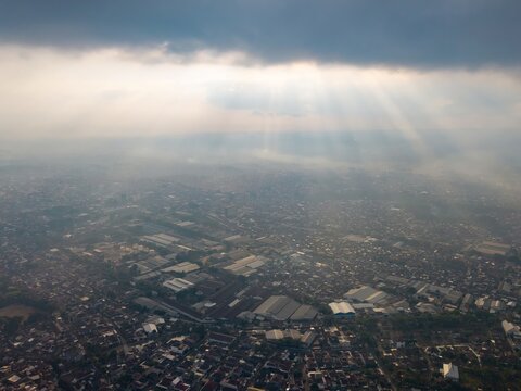 Aerial view of the city of Malang with dense urban rooftops and industrial buildings illuminated by dramatic sun rays breaking through clouds in Malang, East Java, Indonesia.