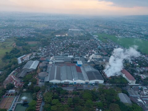 Aerial view of a large sugar factory complex with smoking chimneys, industrial rooftops, and surrounding residential neighborhoods in Malang, East Java, Indonesia.