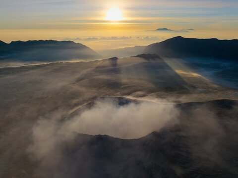 Aerial view of Mount Bromo sunrise with volcanic crater emitting smoke, surrounded by misty mountains and dramatic sun rays at dawn in Tosari, East Java, Indonesia.