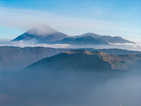 Aerial view of Mount Bromo with Mount Semeru in the background, featuring volcanic craters, rugged ridges, and a sea of clouds under a clear sky in Tosari, East Java, Indonesia.