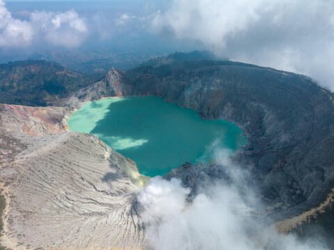 Aerial view of Ijen crater lake with its turquoise water, rugged volcanic slopes, and surrounding clouds in Ijen, East Java, Indonesia.