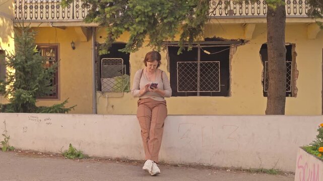 A young female tourist with a backpack rests against an urban parapet and types a text message on her mobile phone on a city street.