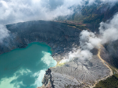 Aerial view of Ijen crater lake with its vibrant turquoise water and sulfurous volcanic smoke rising from the rugged rocky rim in Ijen, East Java, Indonesia.