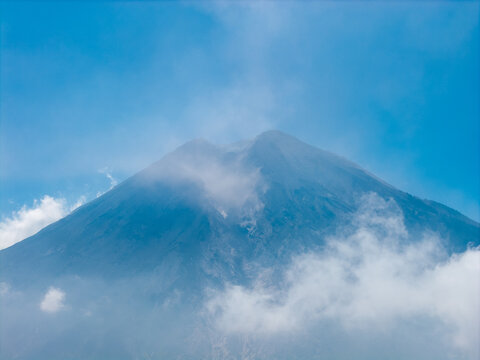 Aerial view of Mount Semeru volcano peak partially covered by clouds under a clear blue sky in Pronojiwo, East Java, Indonesia.