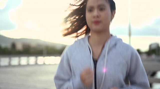 A woman is jogging along a waterfront path in sportswear, with bent arms and a steady running stride. The woman passes a railing while water and distant buildings stretch behind the pavement.