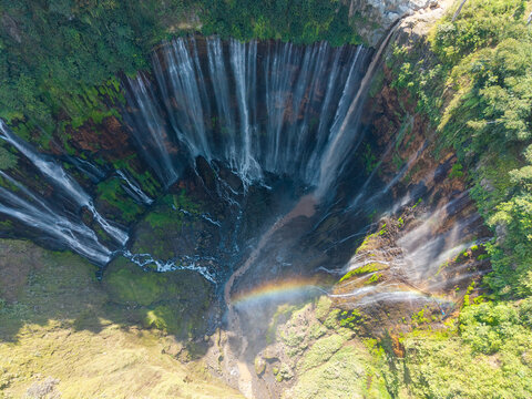 Aerial view of Tumpak Sewu Waterfall featuring cascading streams over a lush green cliffside with a visible rainbow in the mist in Kecamatan Pronojiwo, Jawa Timur, Indonesia.