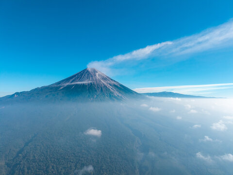 Aerial view of Mount Semeru with a volcanic smoke plume rising above lush forested slopes and a sea of clouds under a clear blue sky in Pasrujambe, East Java, Indonesia.
